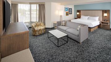 A hotel room with a bed, a sofa, a chair, a TV, a lamp, and a small table.