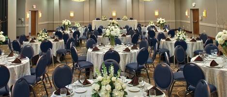 A banquet hall with round tables set for dining, each with white tablecloths, dark blue chairs, and floral centerpieces.