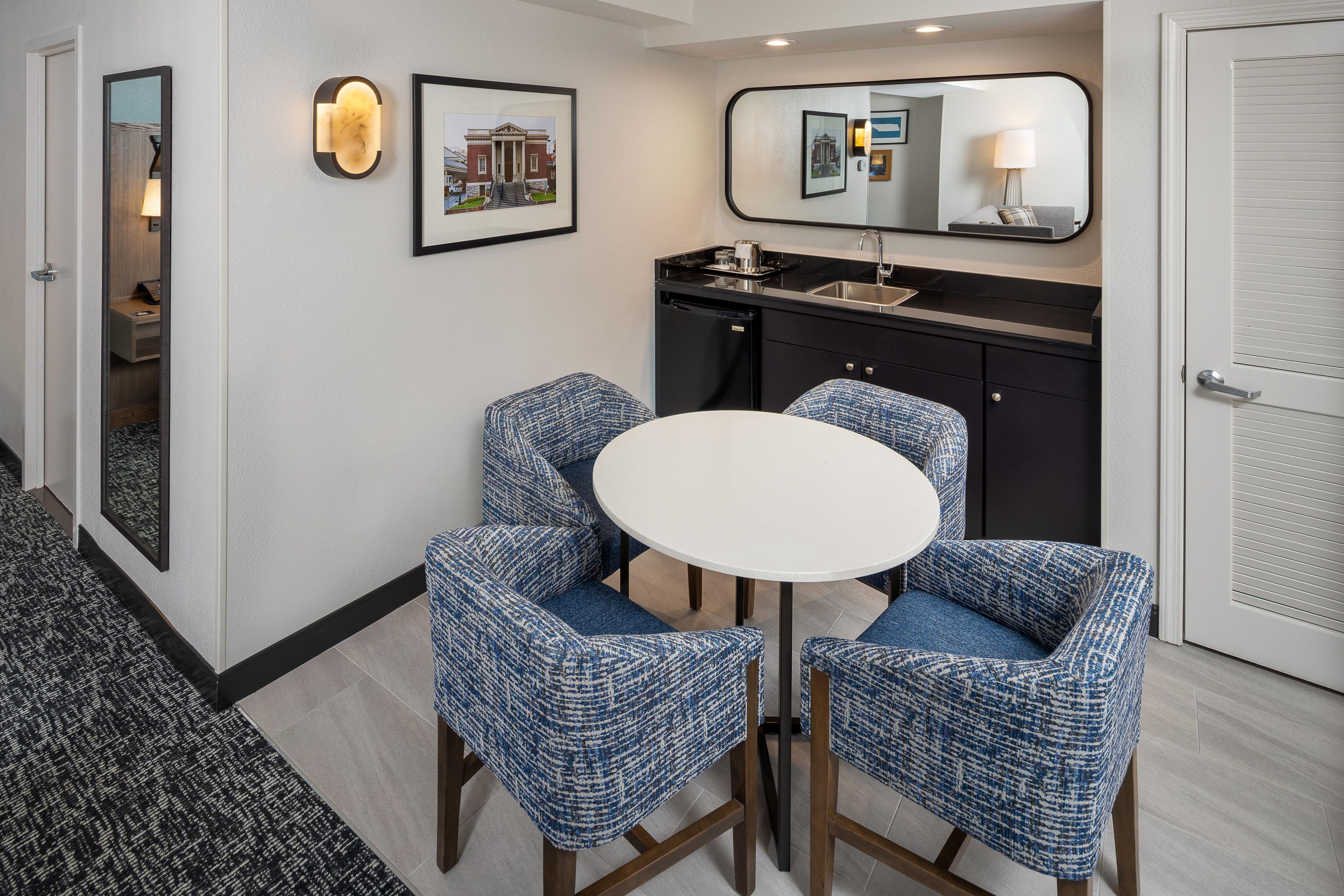 A hotel room with a round table, two blue upholstered chairs, a black minibar, a mirror, and a framed picture on the wall.