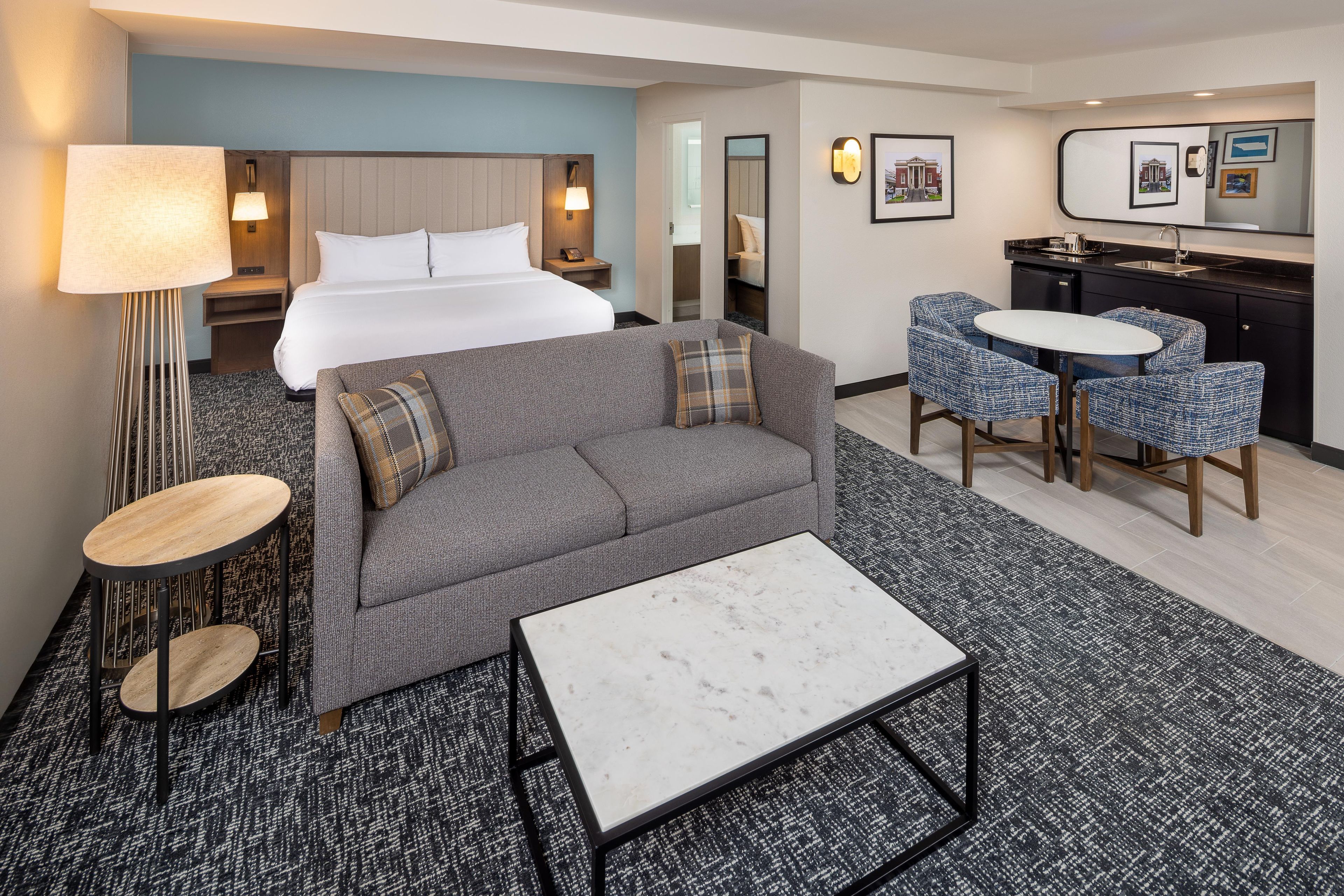 A hotel room with a bed, a sofa, a coffee table, a desk, a chair, a lamp, a mirror, and a small kitchenette.