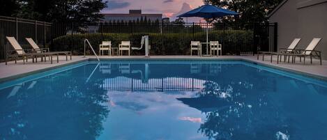 A swimming pool with lounge chairs and a blue umbrella, surrounded by a fence and trees.