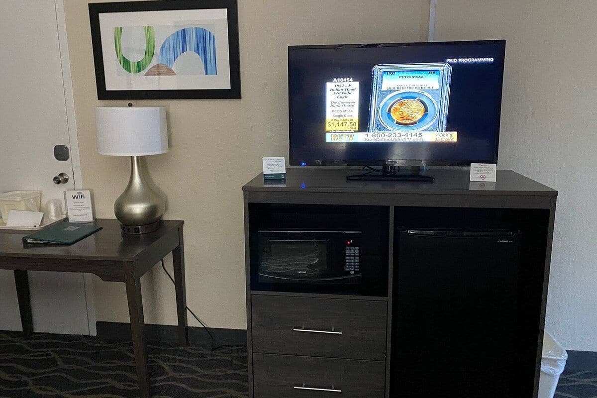 A hotel room with a television displaying a coin grading service, a lamp, a microwave, and a mini-fridge.