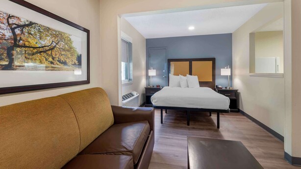 A hotel room with a bed, a brown leather sofa, a framed picture on the wall, a window, and a nightstand with a lamp. - Studio 6 Suites Charlotte, NC - Tyvola Rd/Executive Park (Charlotte)
