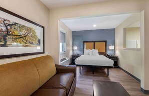 A hotel room with a bed, a brown leather sofa, a framed picture on the wall, a window, and a nightstand with a lamp.