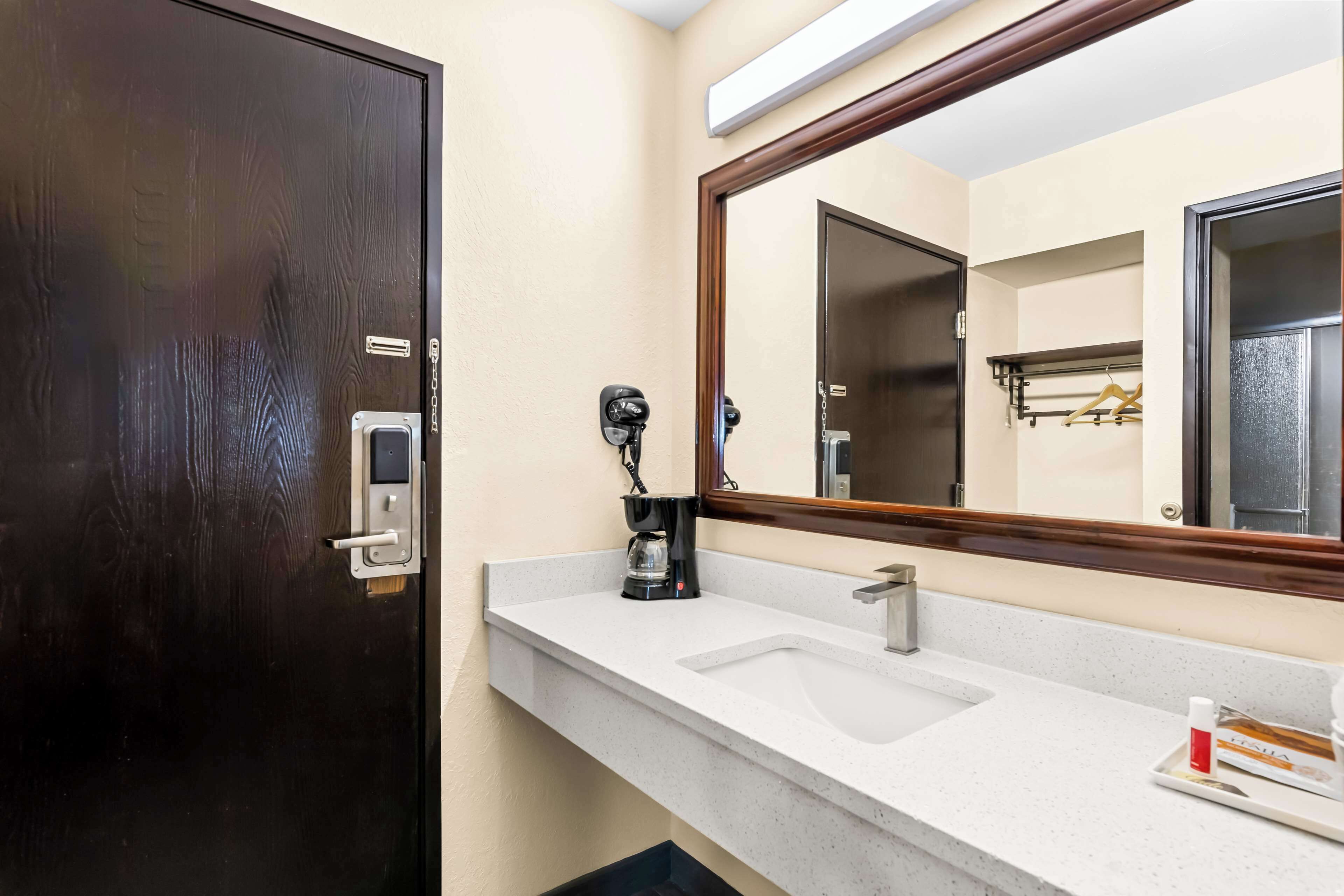 Une salle de bain avec une porte en bois foncé, un lavabo blanc avec un miroir au-dessus, et une machine à café posée sur le comptoir.