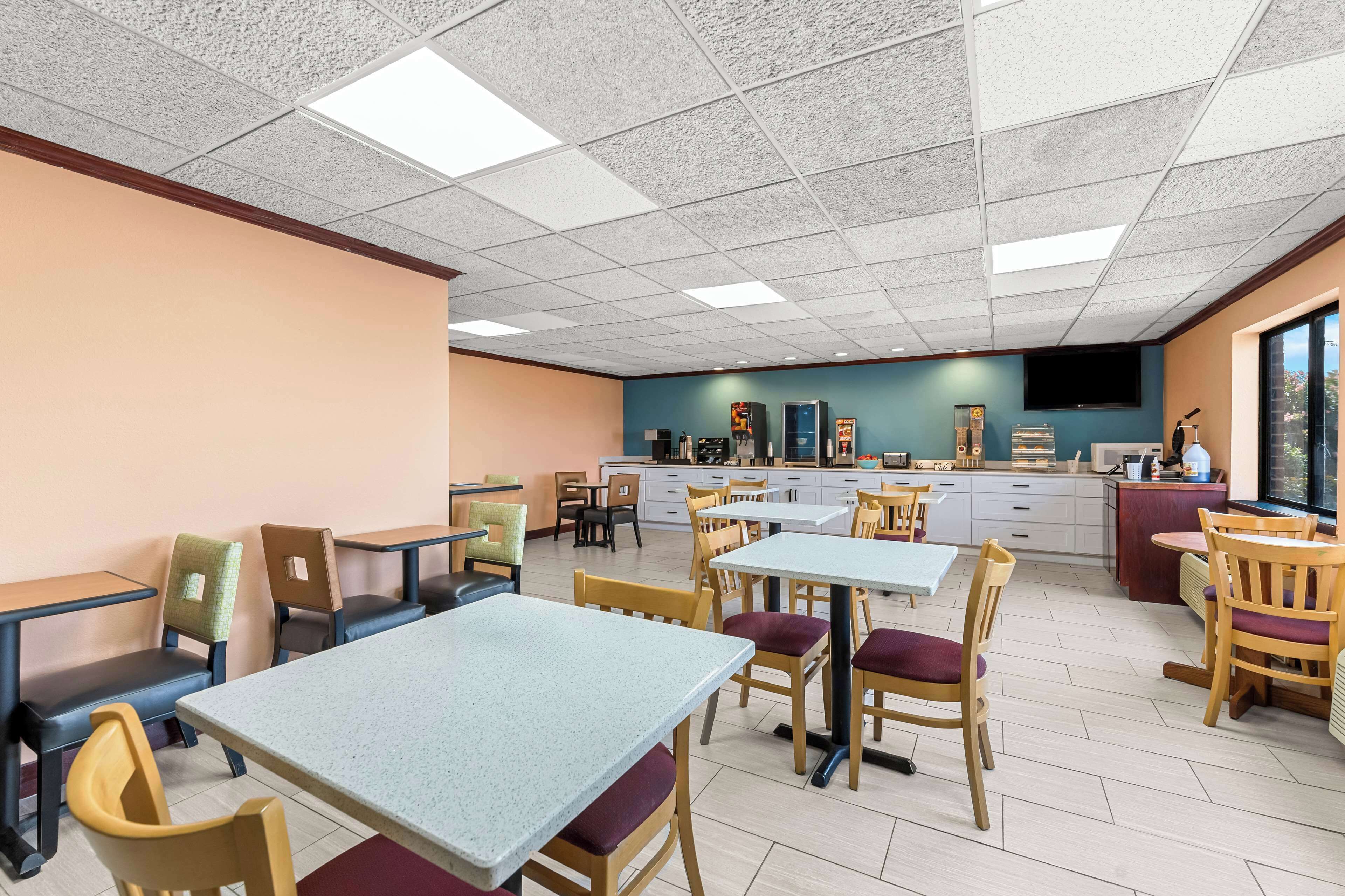 A spacious dining area with multiple tables and chairs, a kitchen area in the background, and a large window providing natural light.