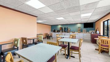 A spacious dining area with multiple tables and chairs, a kitchen area in the background, and a large window providing natural light.