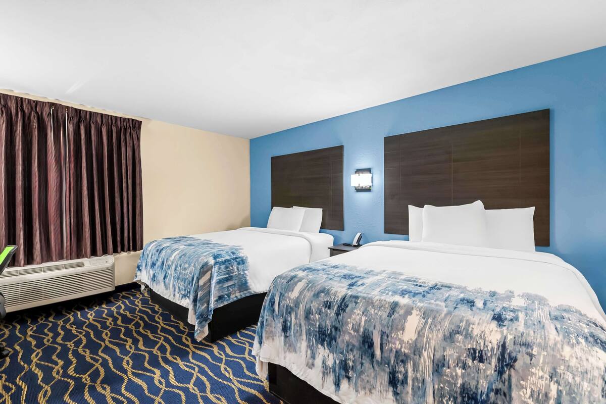 A hotel room with two beds, a blue accent wall, a patterned carpet, and a wall-mounted air conditioner.