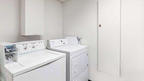 Une buanderie avec deux machines à laver et deux sèche-linge blancs, un meuble de rangement au-dessus des sèche-linge et une porte sur le côté droit.