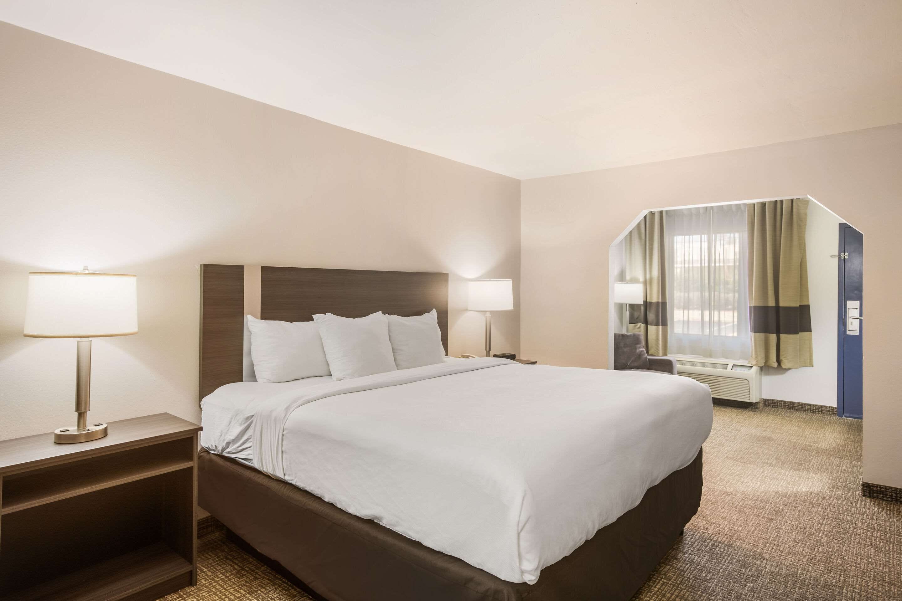 Habitación de hotel con una cama grande, dos mesitas de noche con lámparas, una ventana con cortinas y piso alfombrado.