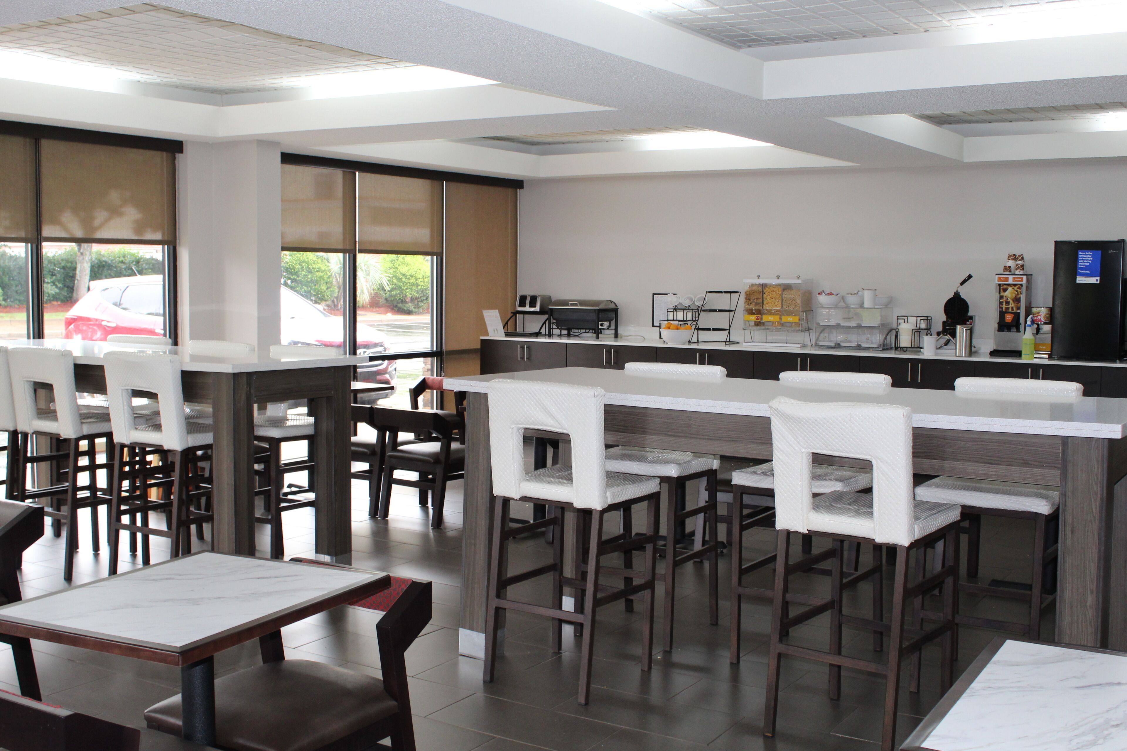 Un restaurant moderne avec des tables et des chaises hautes, un comptoir avec une machine à café et de grandes fenêtres équipées de stores.