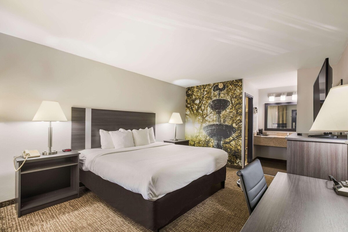 A hotel room with a bed, bedside tables with lamps, a desk with a chair, a mirror, and a wall with a decorative painting.