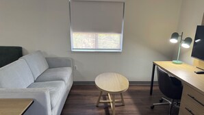 A room with a grey sofa, a round wooden table, a desk with a chair, a standing lamp, and a TV.
