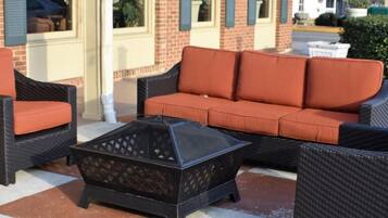 Outdoor wicker furniture with orange cushions, a black fire pit, and a brick building in the background.