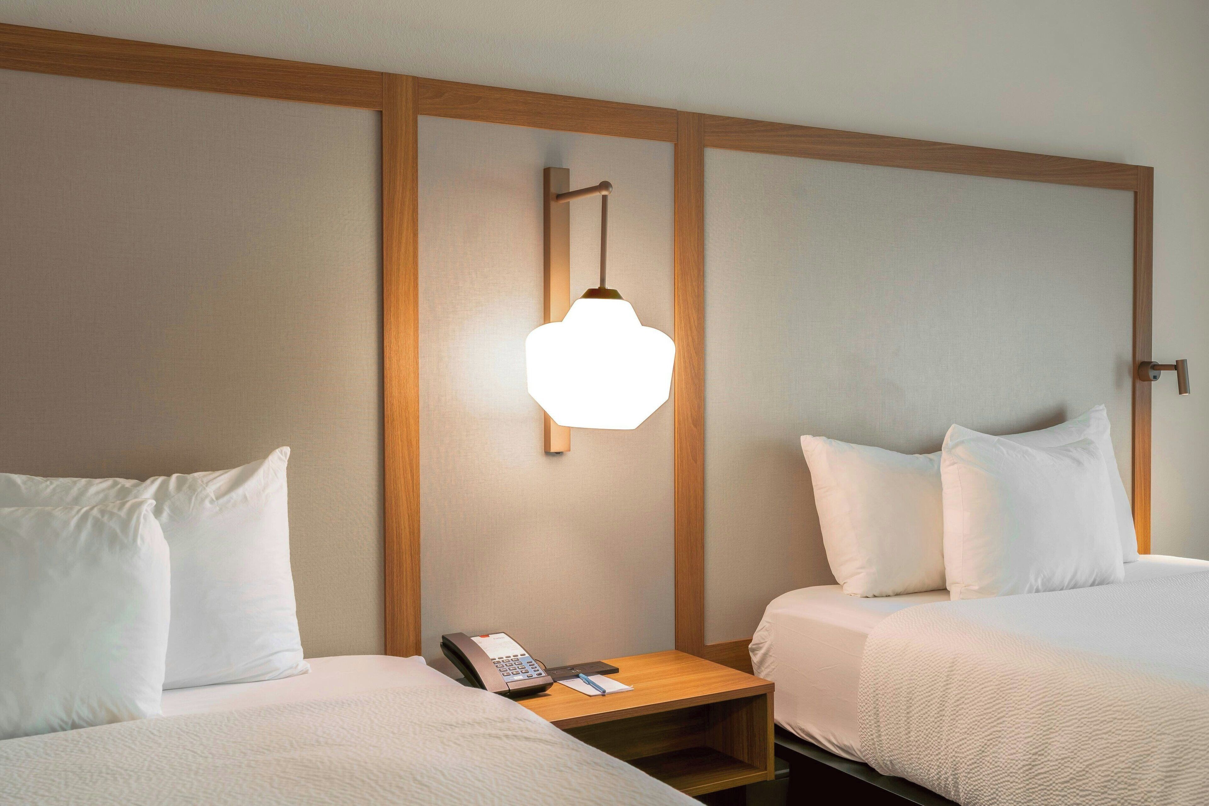 Quarto de hotel com duas camas, uma mesa de cabeceira de madeira com abajur e telefone.