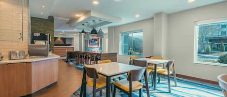 A modern kitchen with a built-in microwave and toaster, a dining area with yellow chairs and a blue patterned rug, and a large window offering a view of the outdoors.