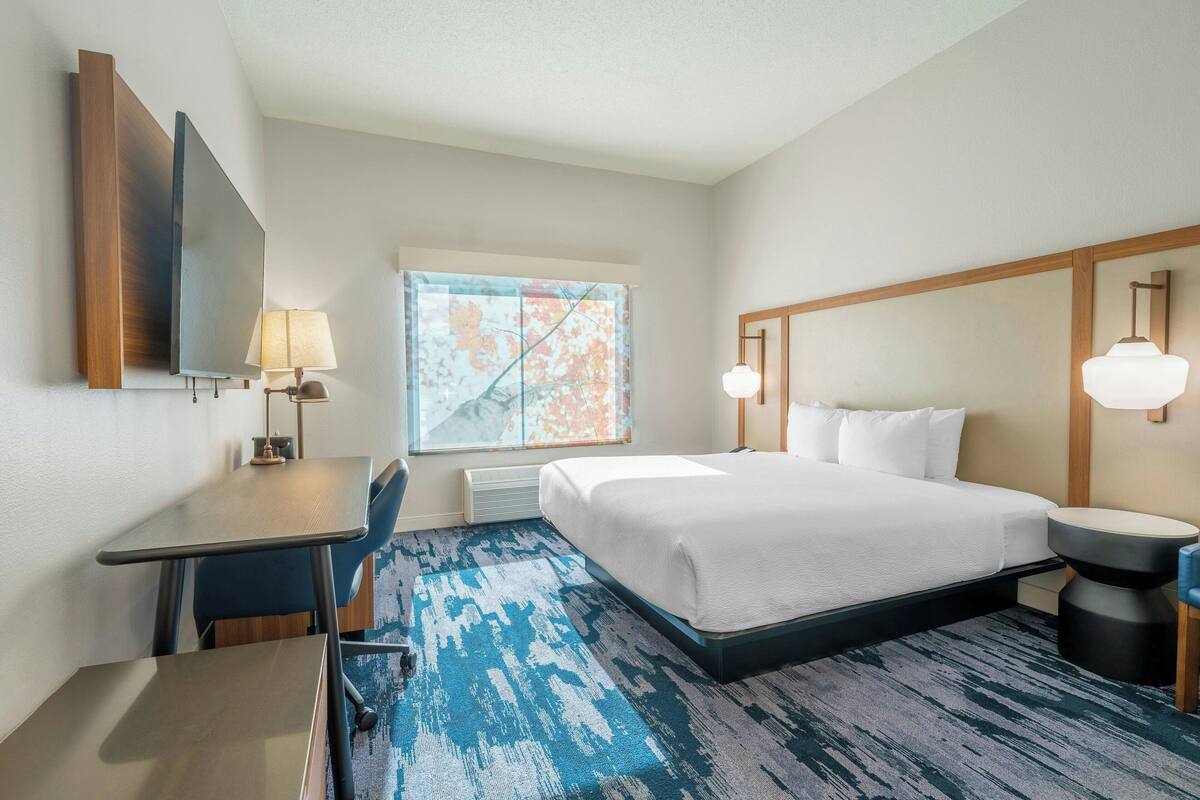 A hotel room with a bed, desk, chair, and a large window.