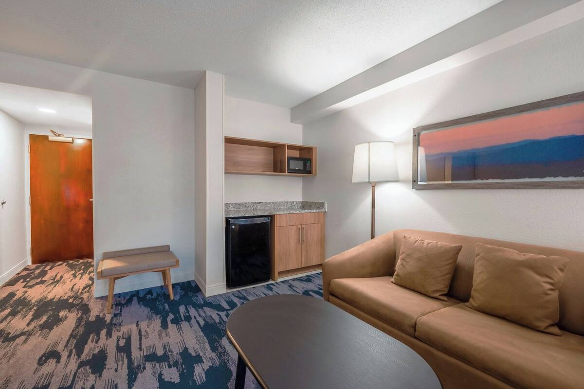 A hotel room with a kitchenette, a sofa, a bench, a small table, a lamp, and a painting on the wall.