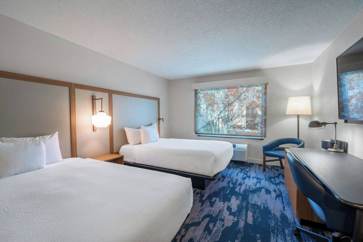 A hotel room with two beds, a desk, a chair, a lamp, and a large window with a view of trees.