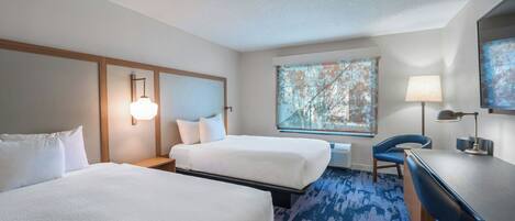 Hotelkamer met twee bedden, een bureau, een stoel, een lamp en een groot raam met uitzicht op bomen.