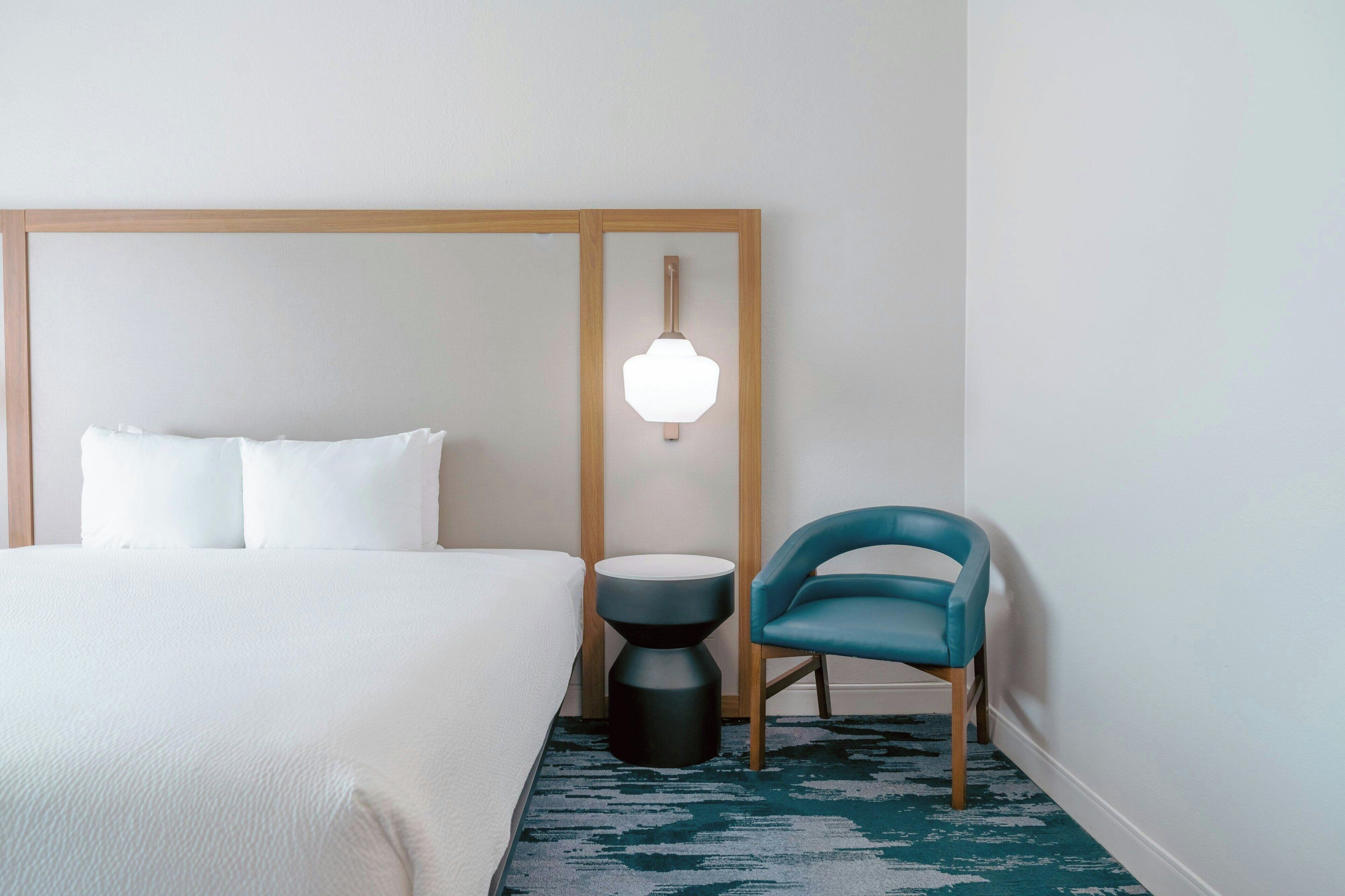 A hotel room with a bed, a round side table, a blue chair, and a wall-mounted light fixture.