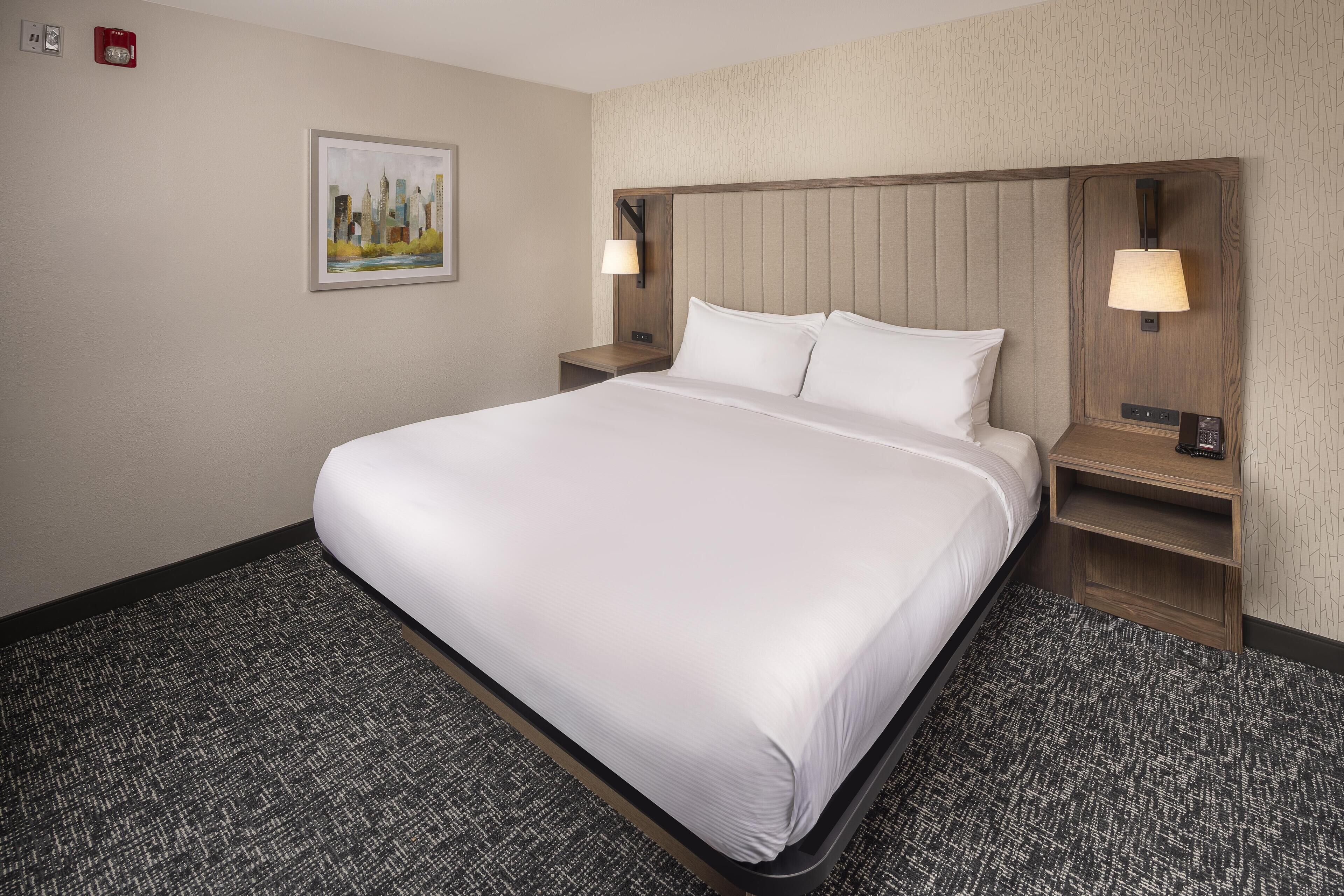 A hotel room with a large bed, two bedside tables with lamps, a painting on the wall, and a carpeted floor.