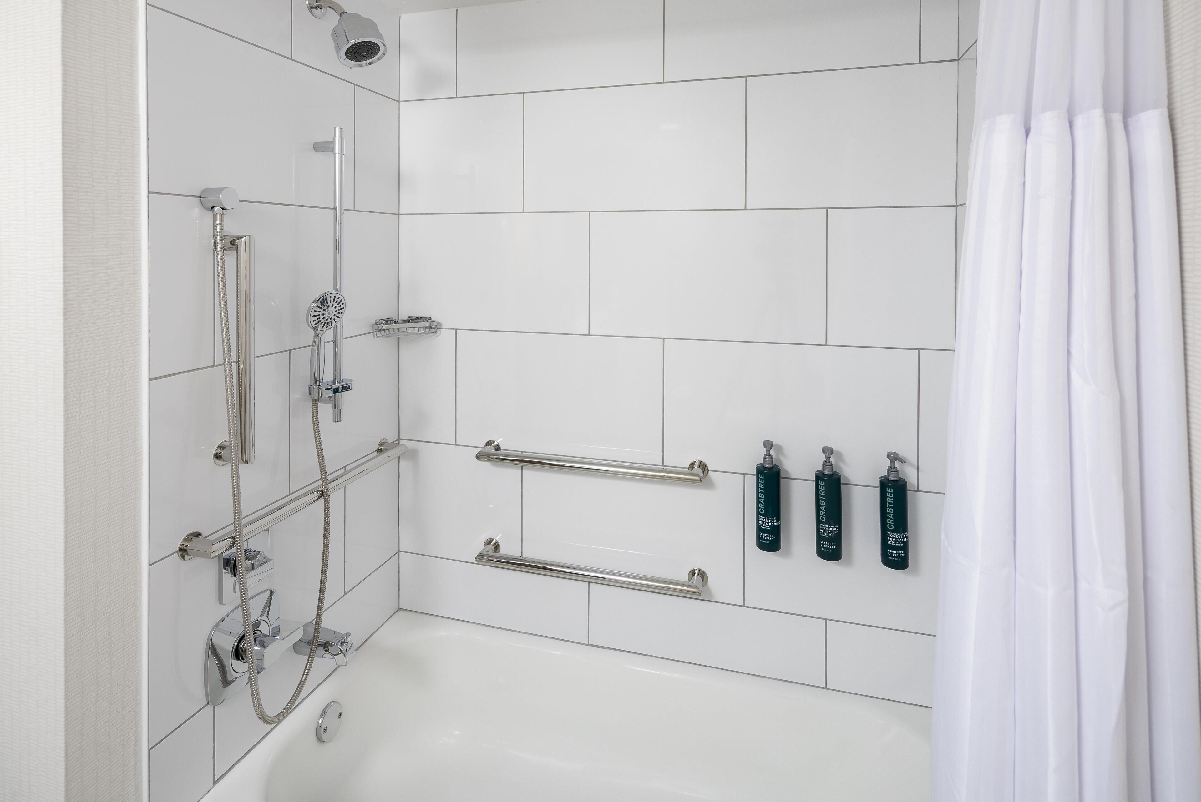 Un baño moderno con azulejos blancos, una bañera, una ducha con cabezal de ducha manual y dos barras de apoyo.
