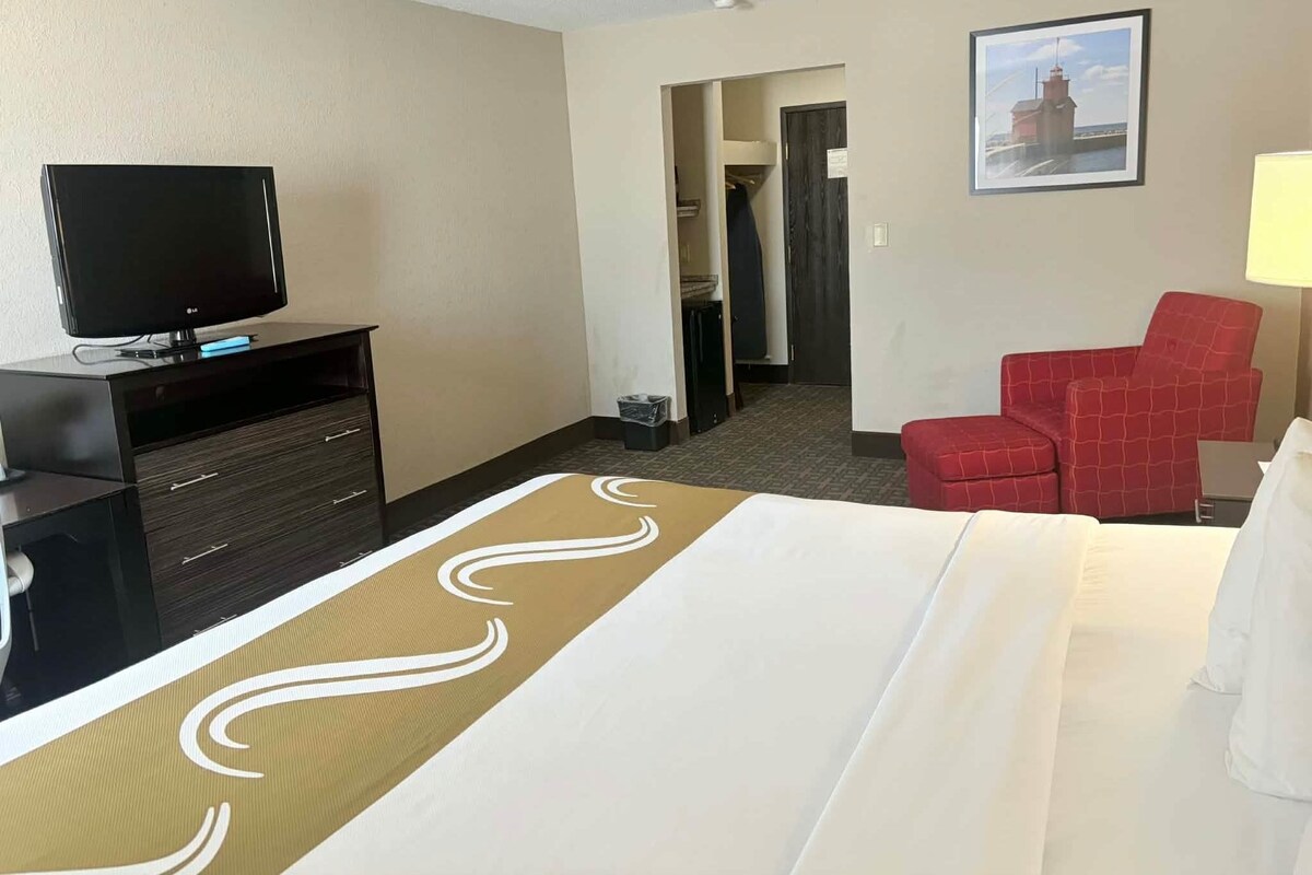 A hotel room with a bed, a TV on a stand, a red chair, and a framed picture on the wall.