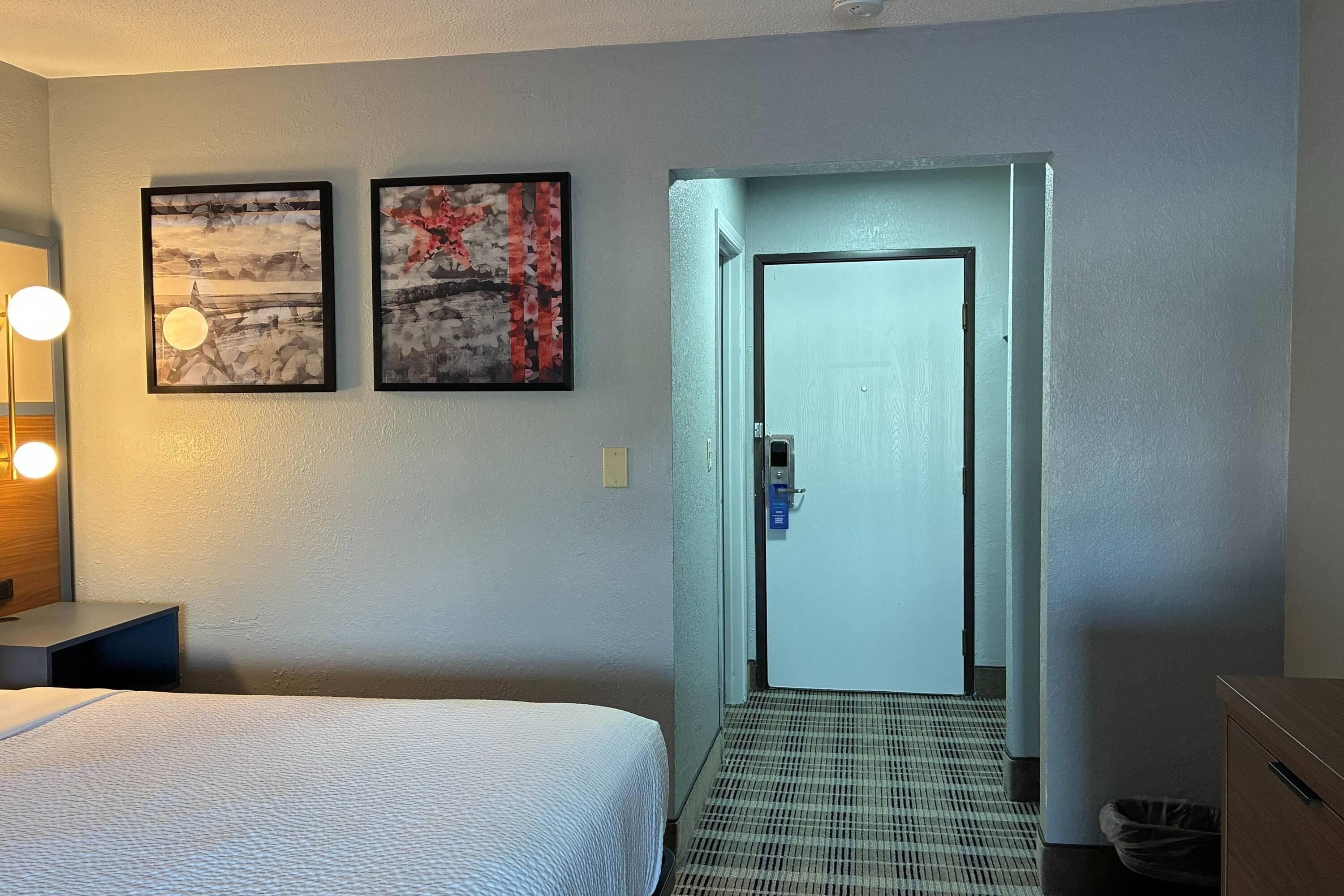 Habitación de hotel con cama, escritorio con lámpara, dos cuadros en la pared y una puerta que da a otra habitación.
