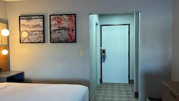 Hotellihuone, jossa on sänky, työpöytä lampun kanssa, kaksi kehystettyä taideteosta seinällä ja ovi, joka johtaa toiseen huoneeseen.