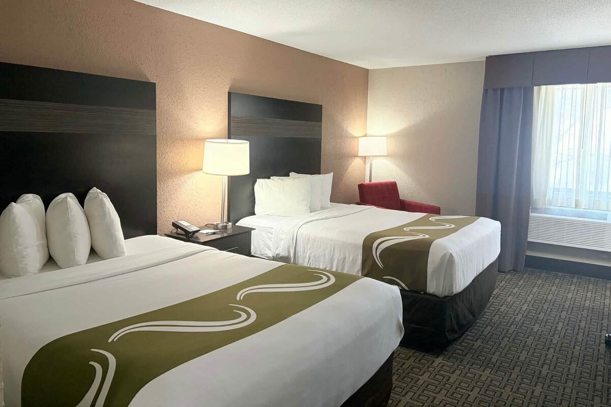 A hotel room with two beds, a nightstand, a red chair, and a window with curtains.