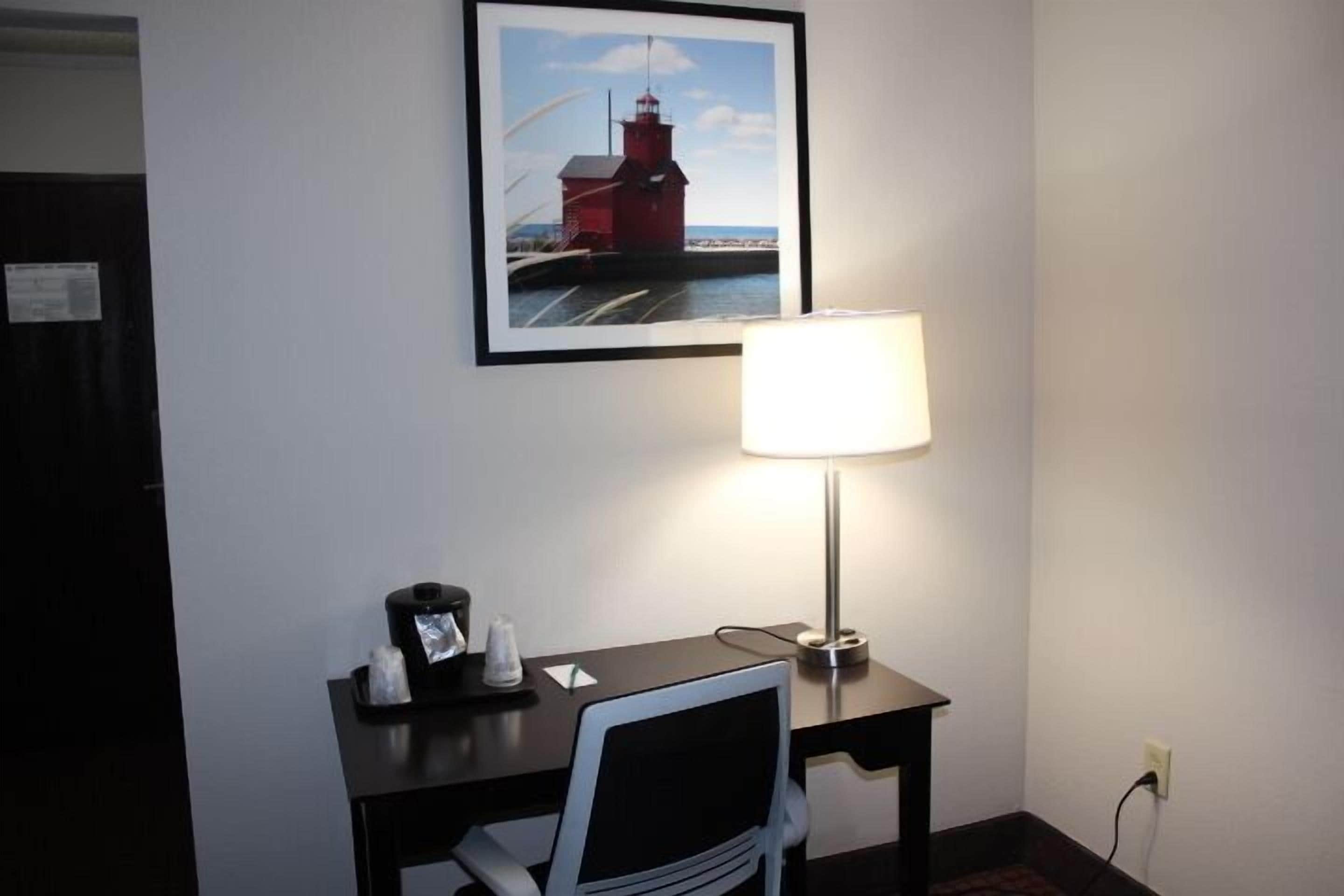 Una habitación de hotel con escritorio, una silla, una lámpara, un cuadro enmarco de un faro y un reloj pequeño.