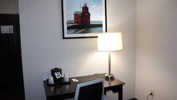 Una habitación de hotel con escritorio, una silla, una lámpara, un cuadro enmarco de un faro y un reloj pequeño.