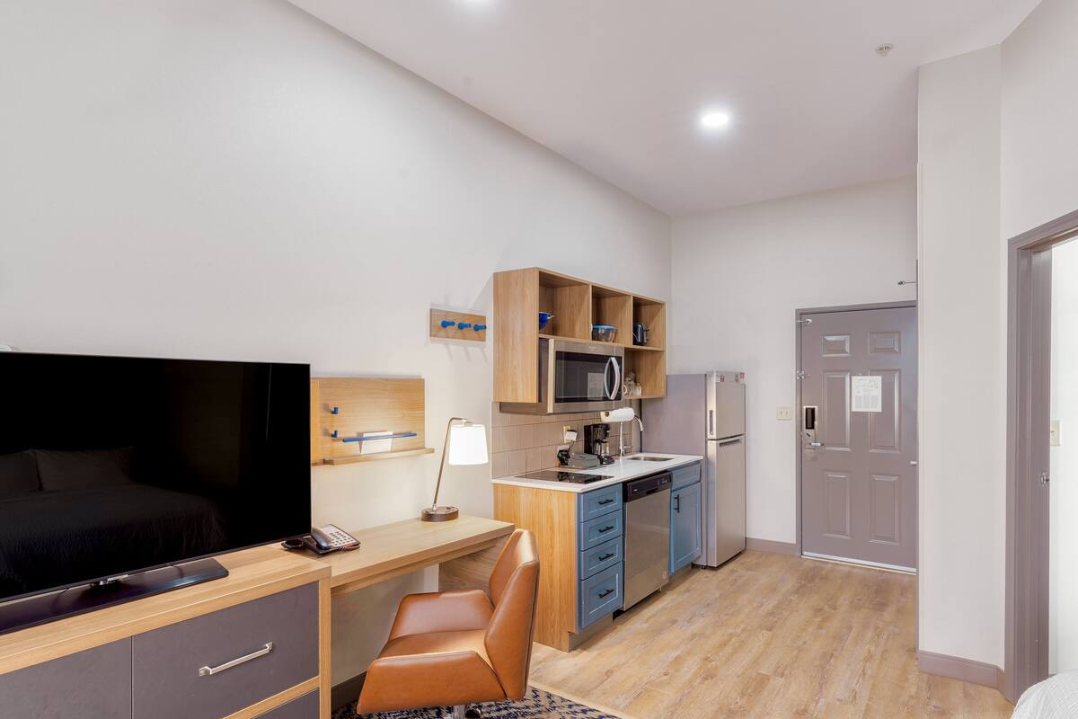 A compact hotel room with a bed, a desk, a chair, a television, a microwave, a refrigerator, and a small kitchenette.