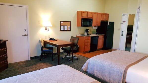 A hotel room with two beds, a desk with a chair, a kitchenette, and a door.
