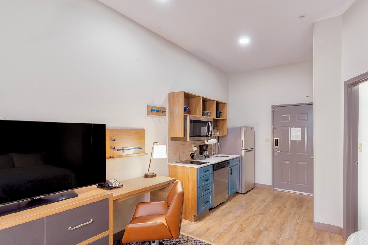 Habitación de hotel pequeña con cama, escritorio, silla, televisor, microondas, refrigerador y puerta.