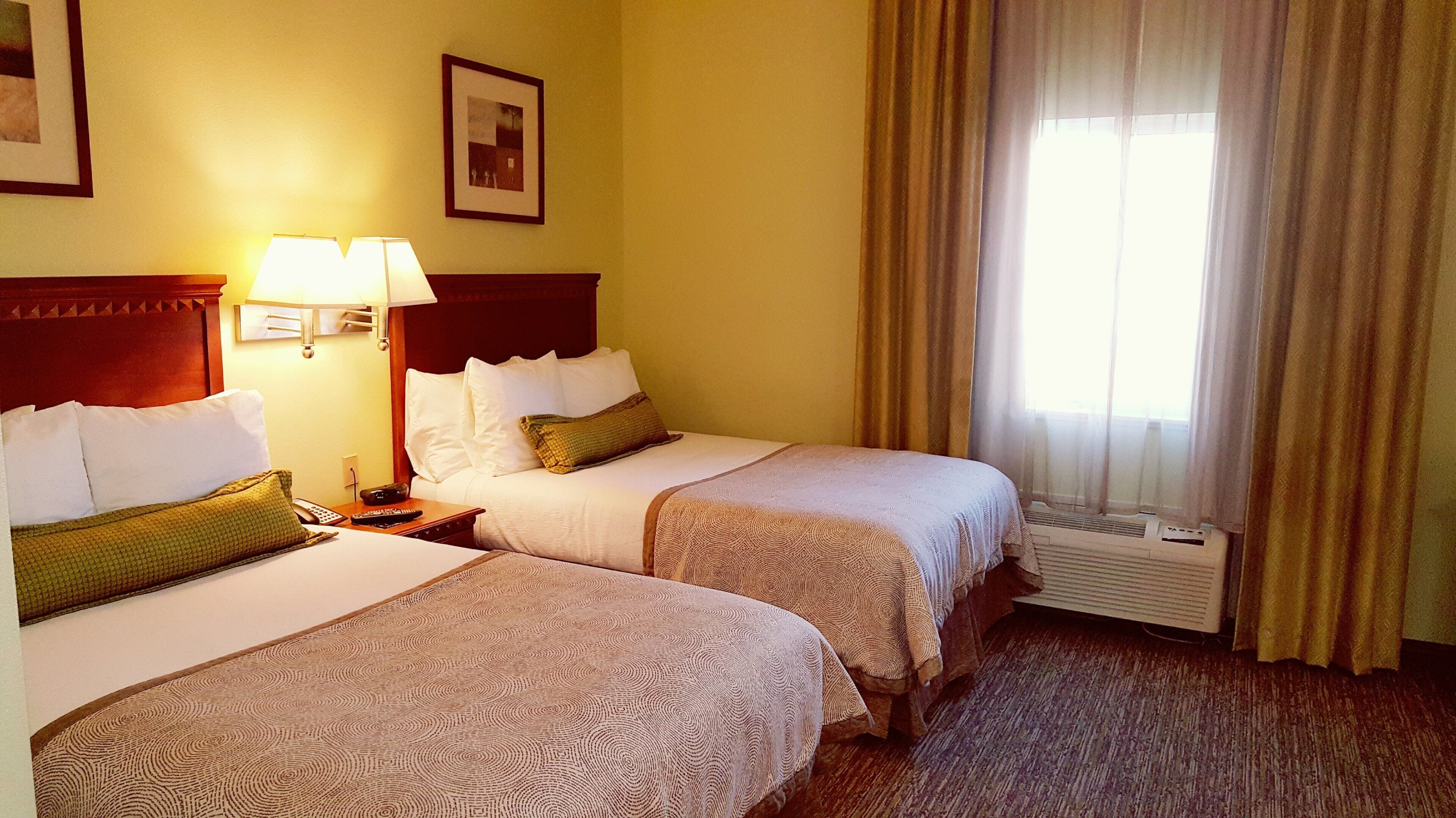 A hotel room with two beds, a nightstand with a lamp, a framed picture on the wall, and a window with curtains.