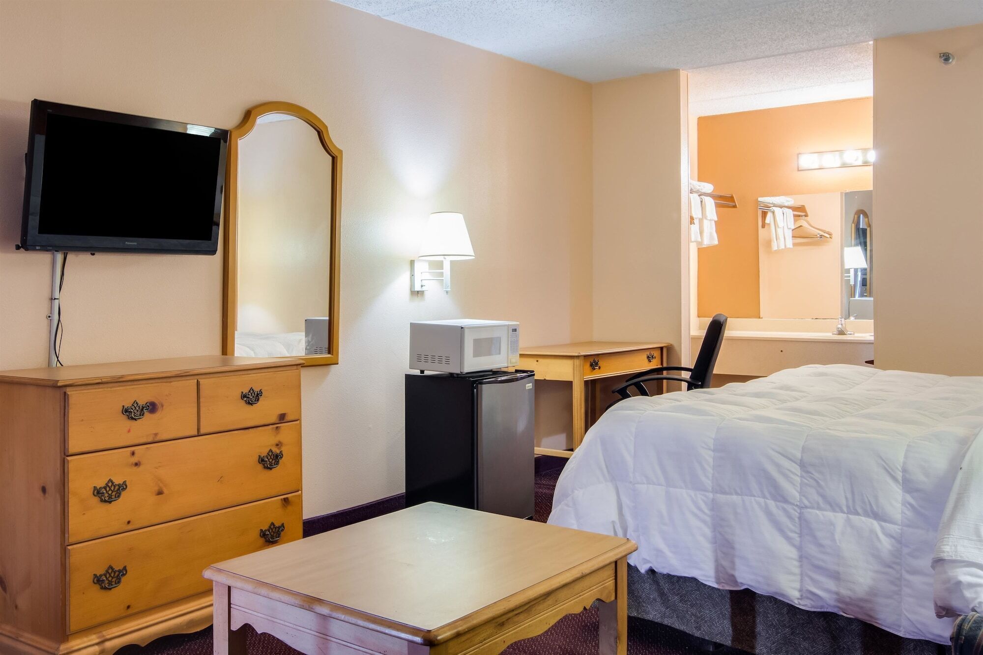 A hotel room with a bed, a wooden dresser, a desk with a computer and a microwave, a TV mounted on the wall, and a bathroom visible through an open door.