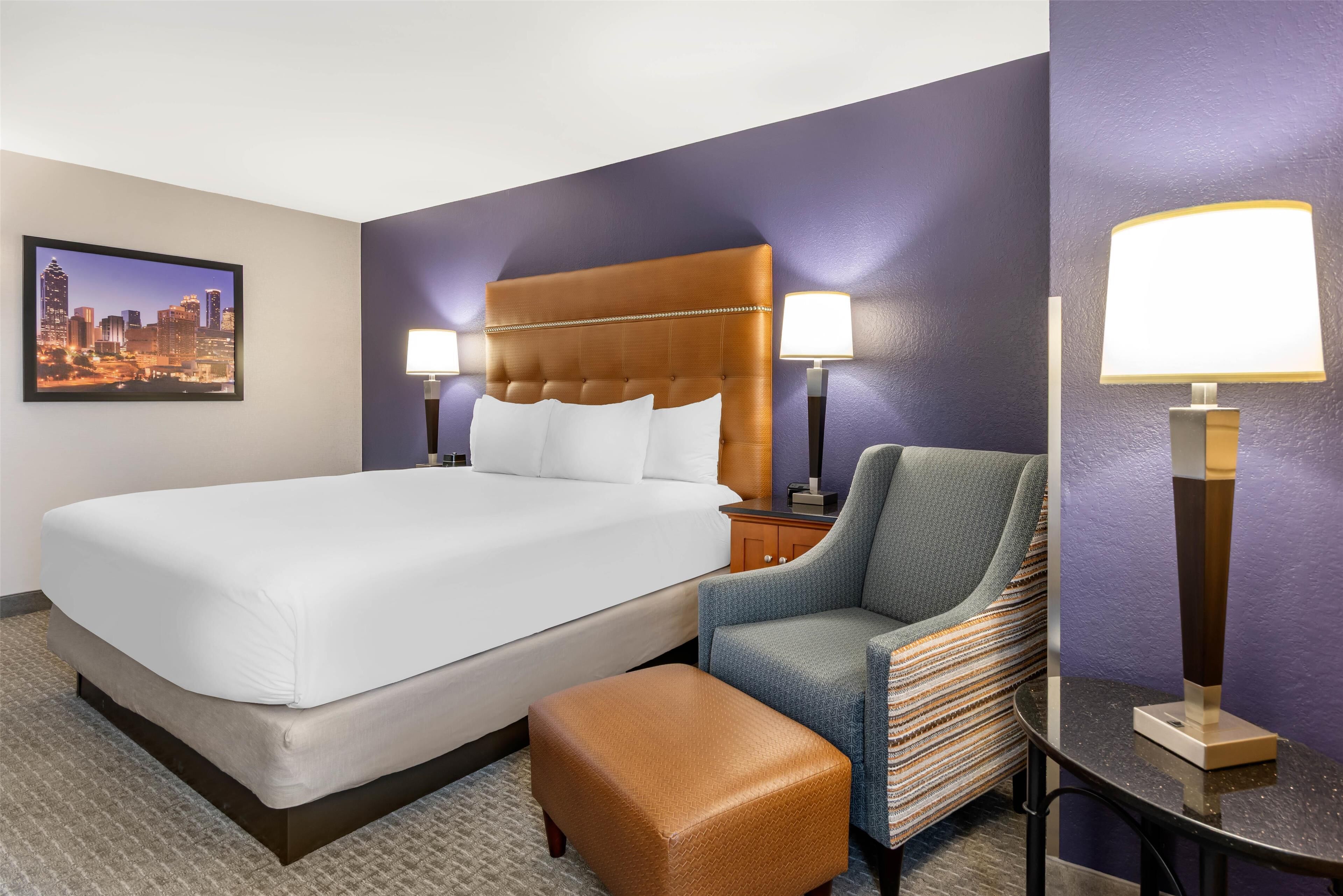 A hotel room with a large bed, a chair, a small table, and two wall-mounted lamps.