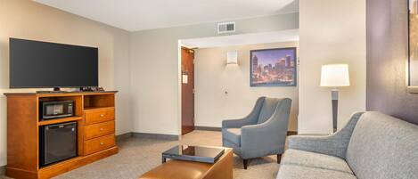 A hotel room with a flat-screen TV, a microwave, a mini-fridge, a sofa, a chair, and a coffee table.