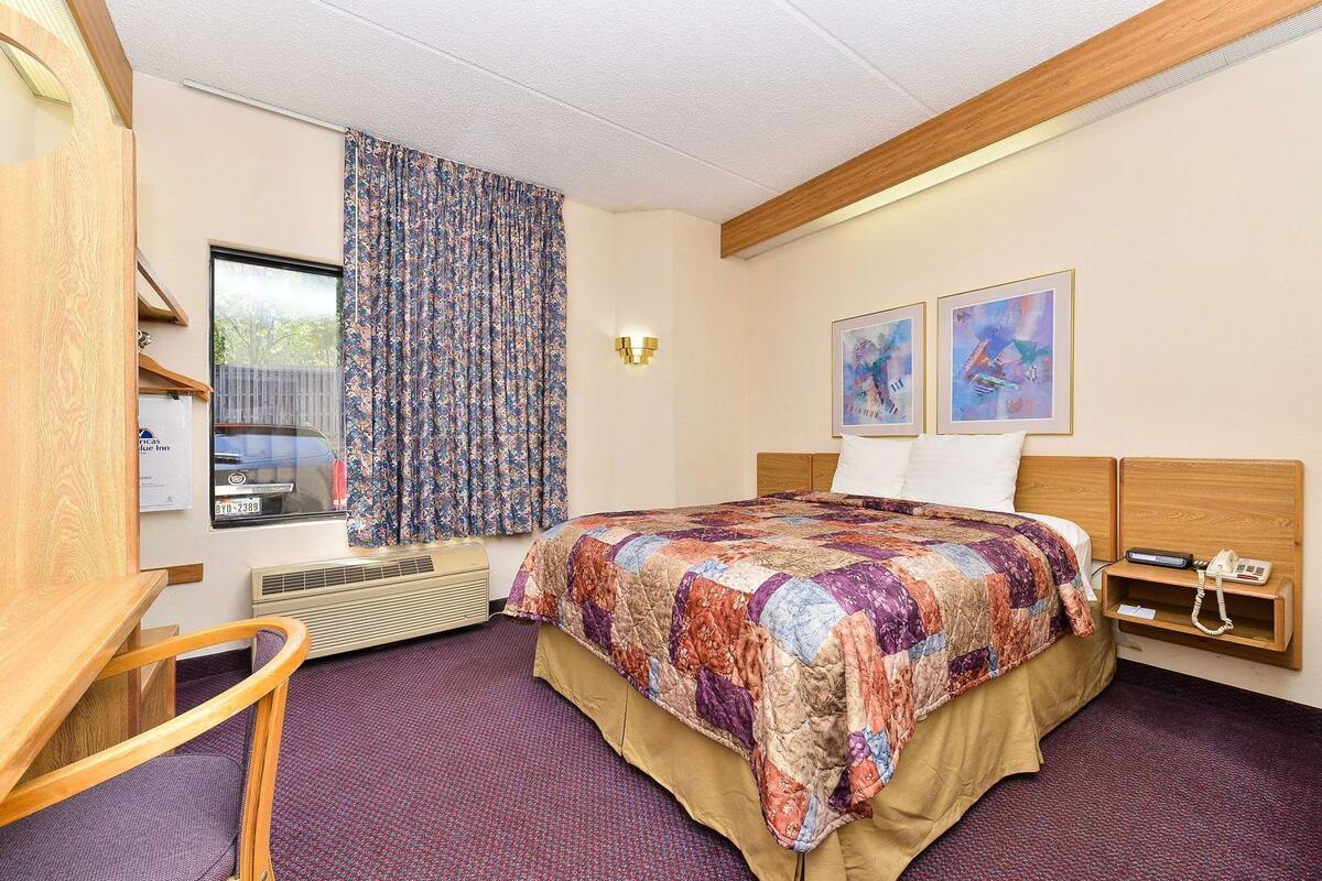 A hotel room with a bed, a desk, a chair, a window with curtains, a wall-mounted heater, and two framed pictures.