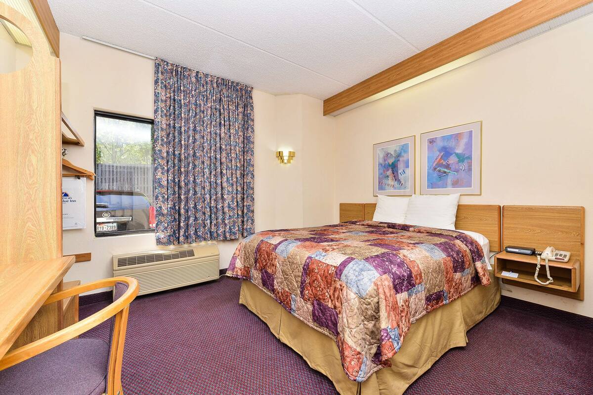 A hotel room with a large bed, a desk, a chair, a window with curtains, and two framed artworks on the wall.