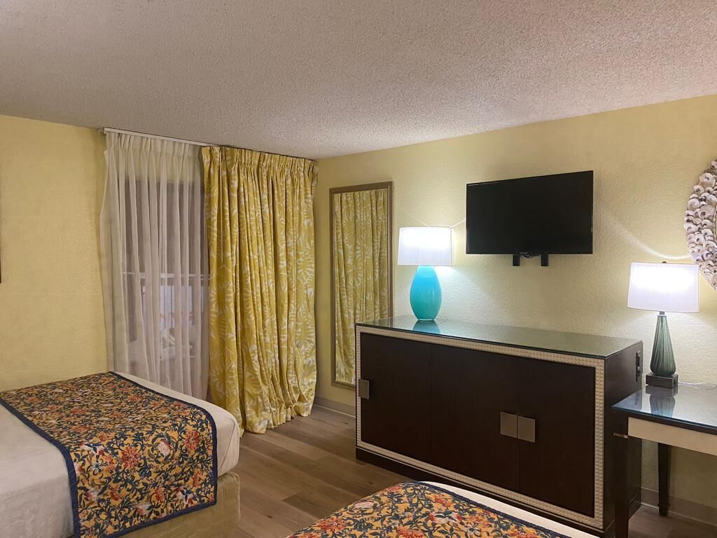 Una habitación de hotel con dos camas, un televisor, un escritorio con lámpara y un ventanal con cortinas.