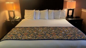 Ein Hotelzimmer mit einem großen Bett, weißen Laken, einer floralen Tagesdecke, zwei Nachttischlampen, einem Telefon auf dem Nachttisch und einer digitalen Uhr.