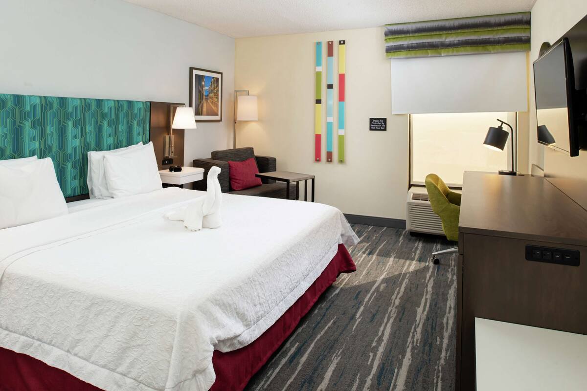 A hotel room with a large bed, a desk, a chair, a TV, and a wall-mounted air conditioner.