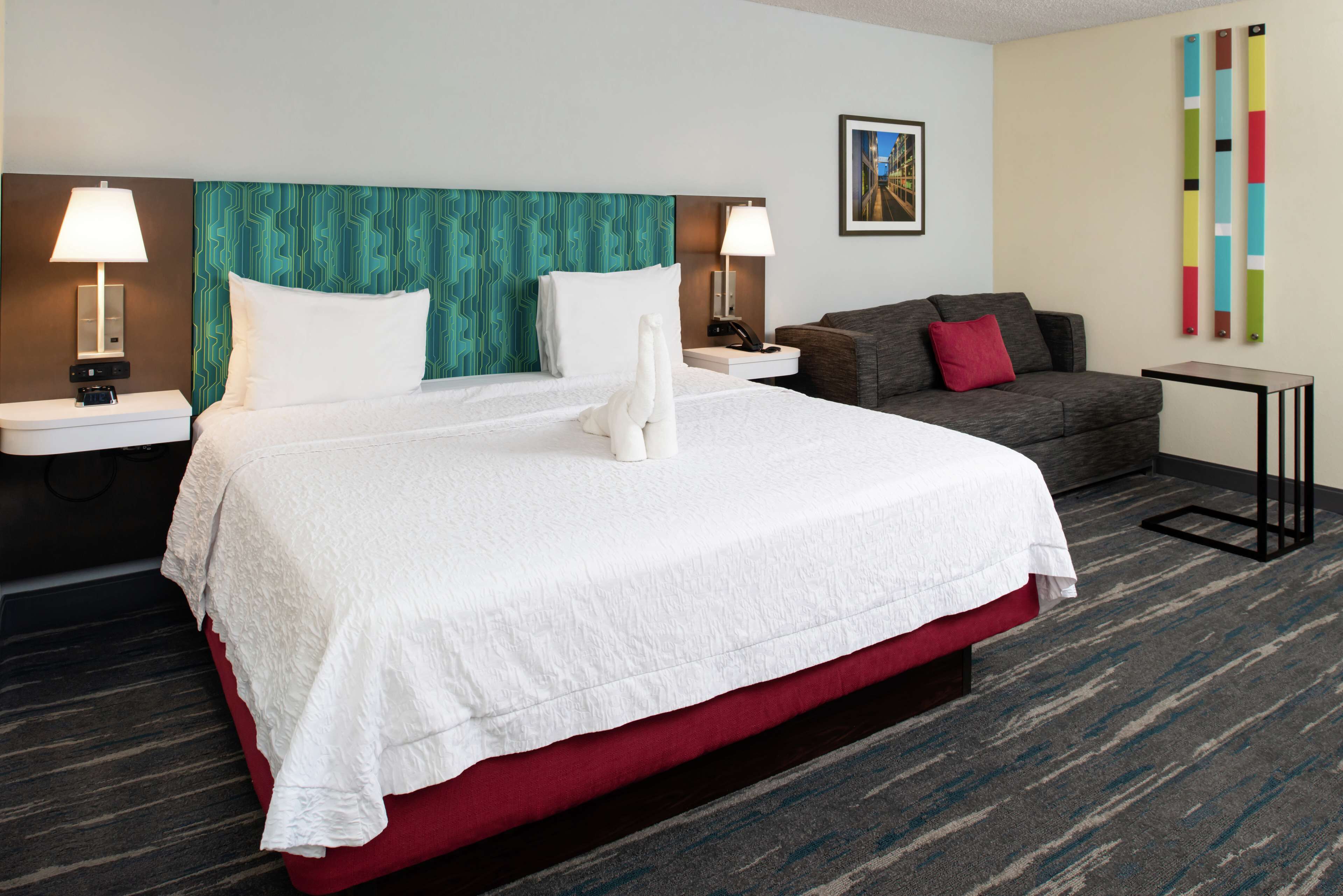 A hotel room with a large bed, two bedside tables with lamps, a gray sofa, and a small table.
