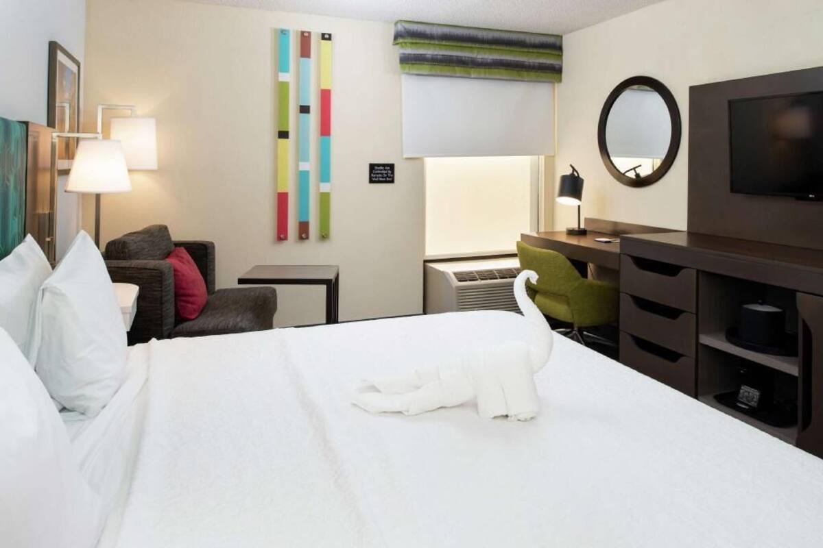 A hotel room with a bed, a desk, a chair, a TV, and a wall with colorful decor.