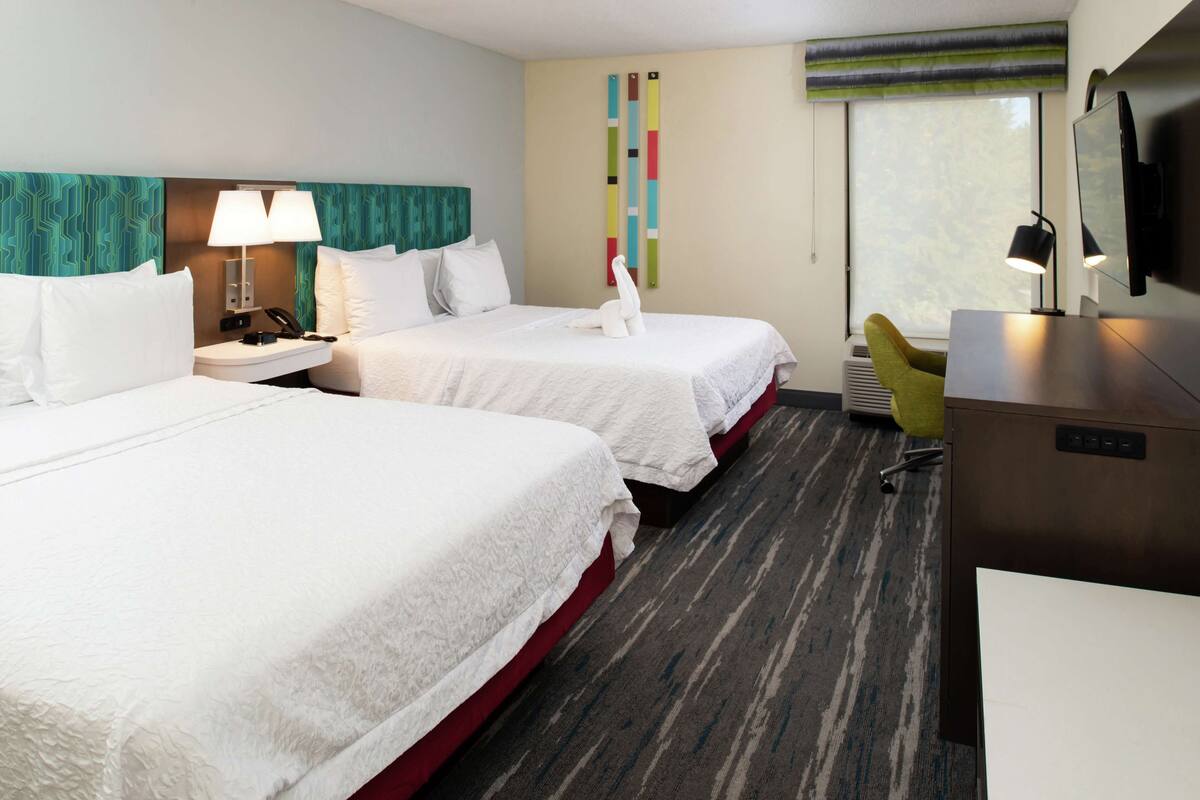 A hotel room with two beds, a desk, a chair, a TV, and a window with a view.