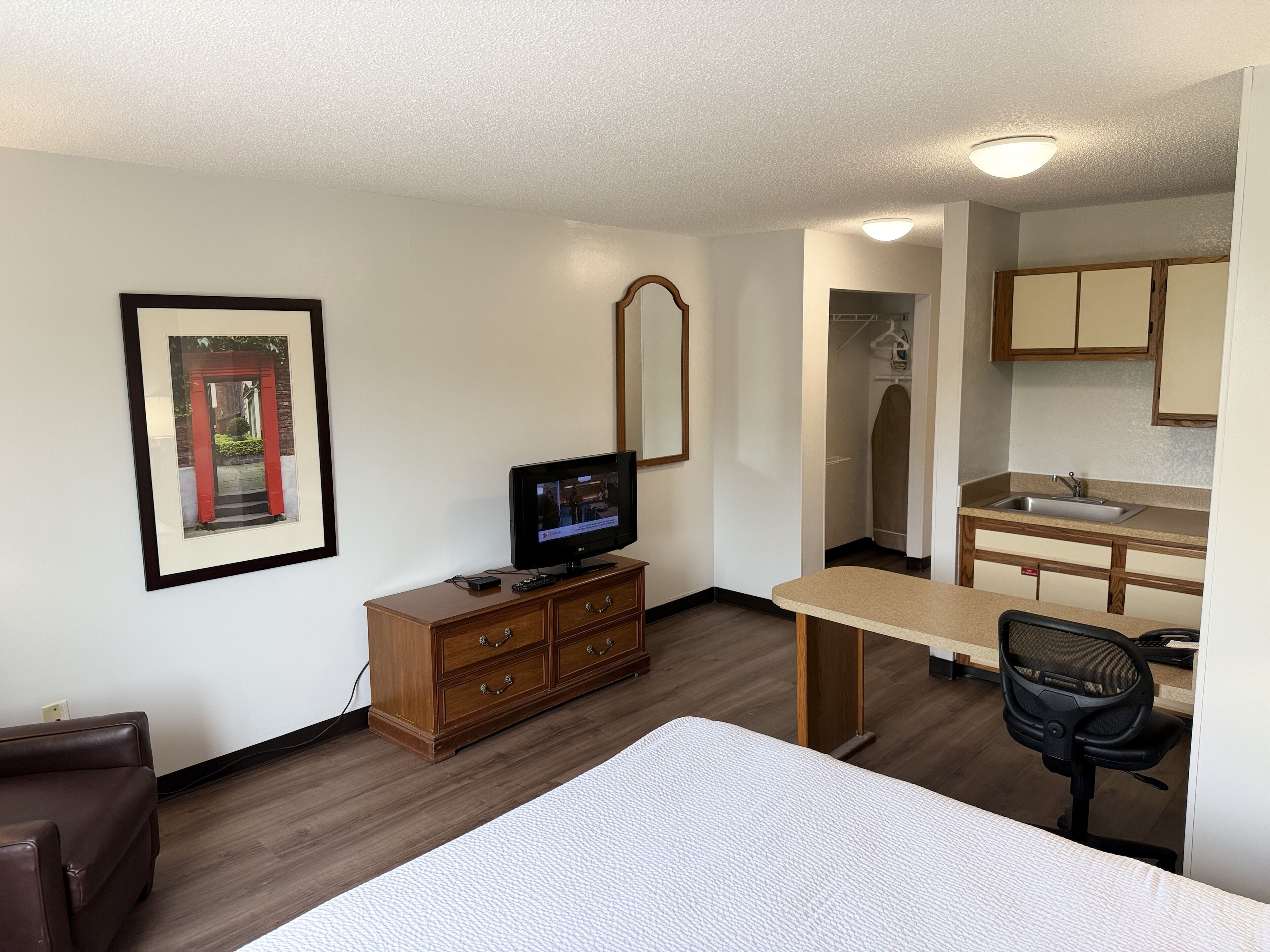 A hotel room with a bed, a TV on a wooden dresser, a desk with a chair, a kitchen area with a sink, and a mirror on the wall.