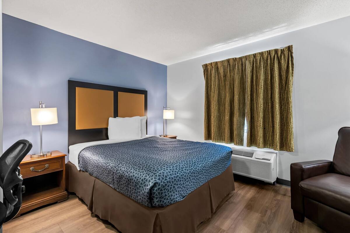 Hotellihuone, jossa on sänky, yöpöydällä olevat lamput, työpöytä, tuoli ja ikkuna, jossa on verhot.